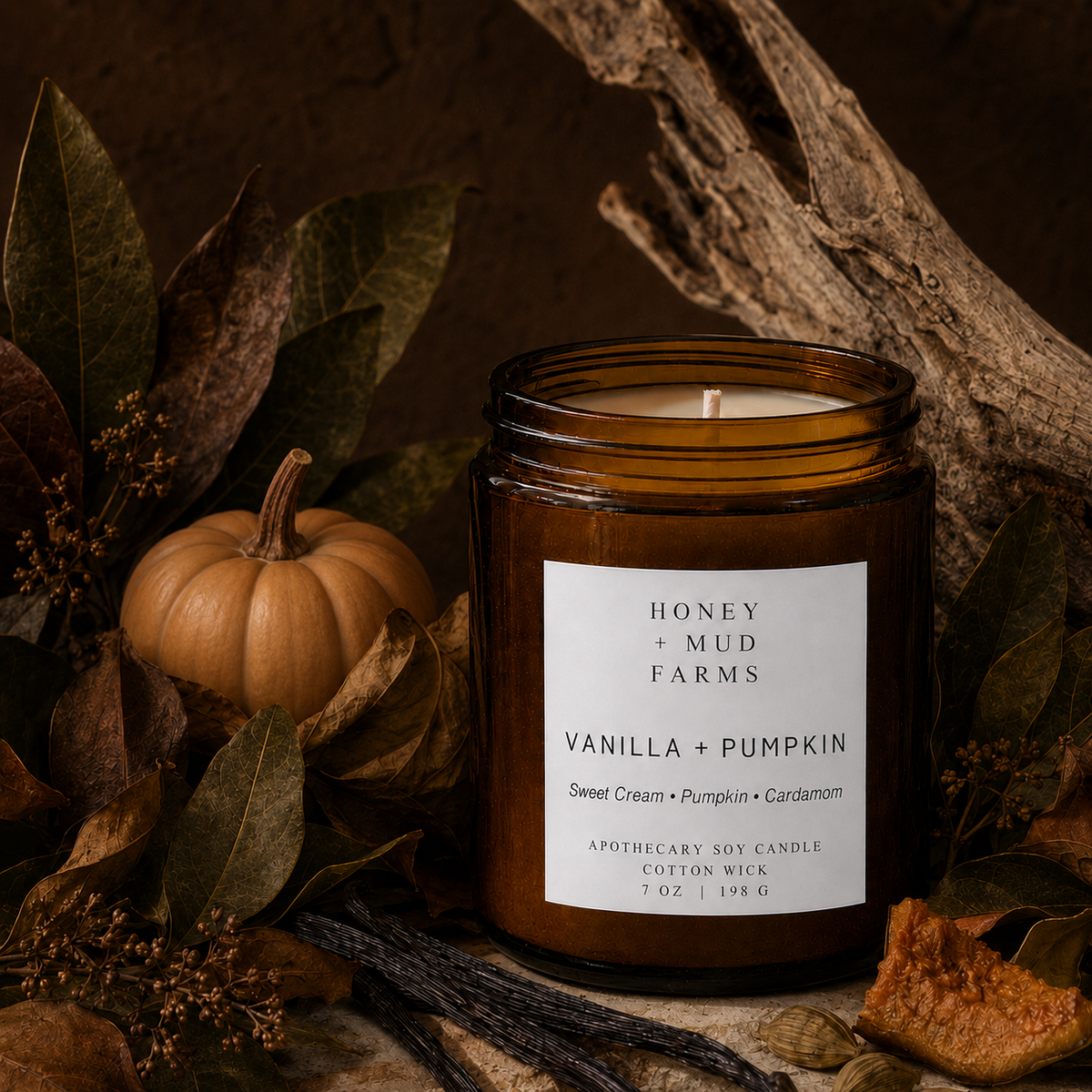 An amber glass jar candle labeled Honey + Mud Farms Vanilla + Pumpkin, styled with autumn leaves, dried grass, a small pumpkin, and driftwood on a dark rustic background.