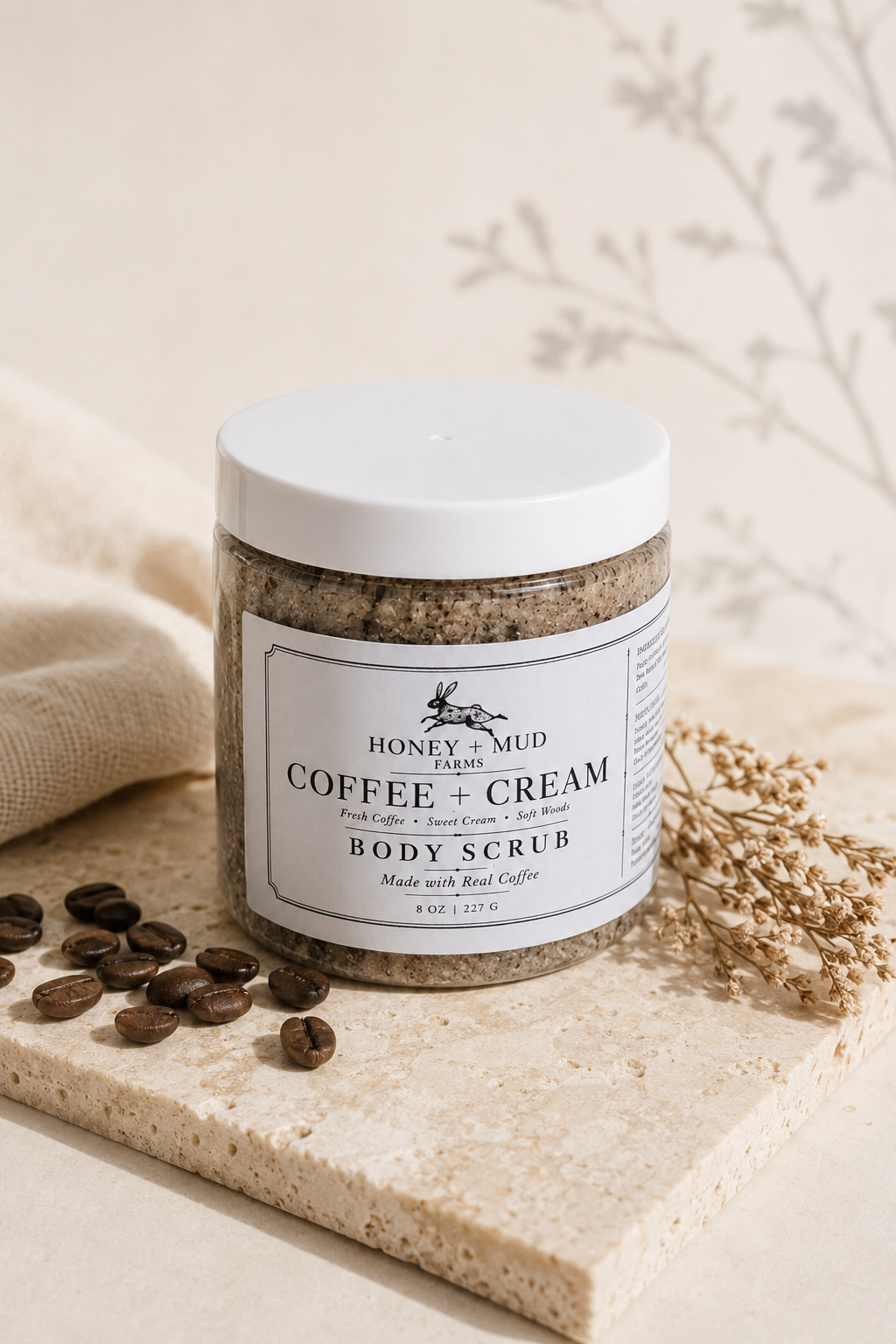 A jar of Honey + Mud Coffee + Cream Body Scrub sits on a stone slab with scattered coffee beans and dried flowers. The clear jar shows a textured coffee scrub inside, with a white lid and a white label featuring black text.