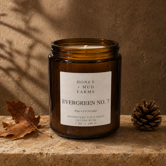 An amber glass jar candle labeled 'Evergreen No. 7' by Honey + Mud Farms, styled on a stone surface with a dry leaf and a pinecone against a textured brown background.