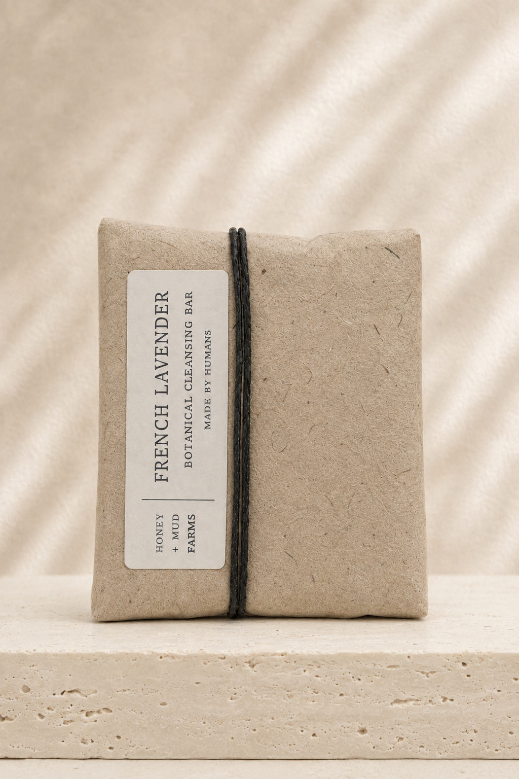 A wrapped natural paper cleansing bar labeled 'French Lavender' by Lavender & Mud Farms, tied with a black string, set on a textured stone surface against a warm beige background with soft leaf shadows.