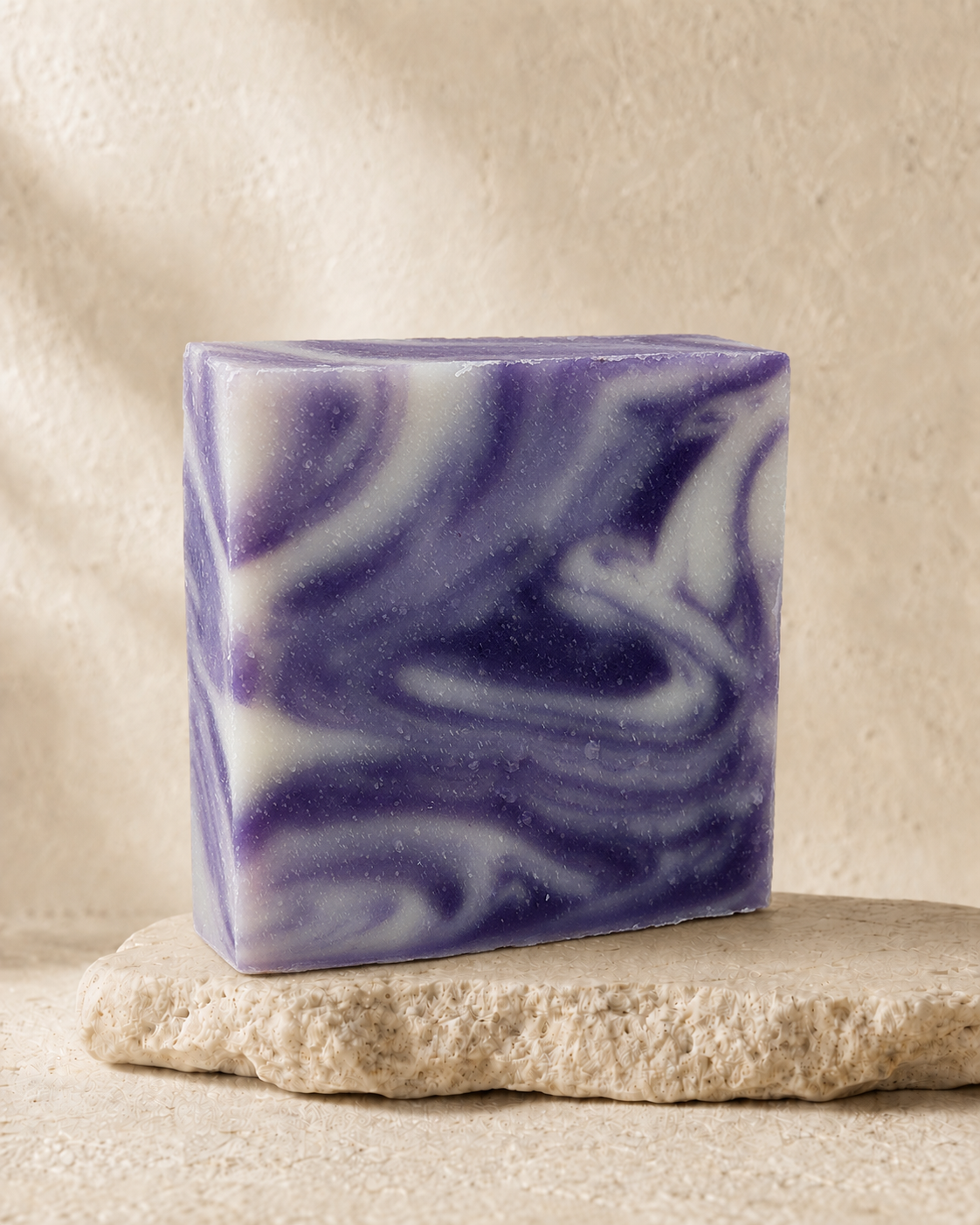 A rectangular bar of handmade soap with a marbled swirl pattern of purple and white, resting on a textured beige stone slab against a neutral beige background.