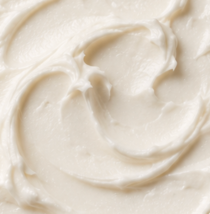 A close-up texture shot of a thick, creamy off-white cosmetic cream with soft swirls and folds.