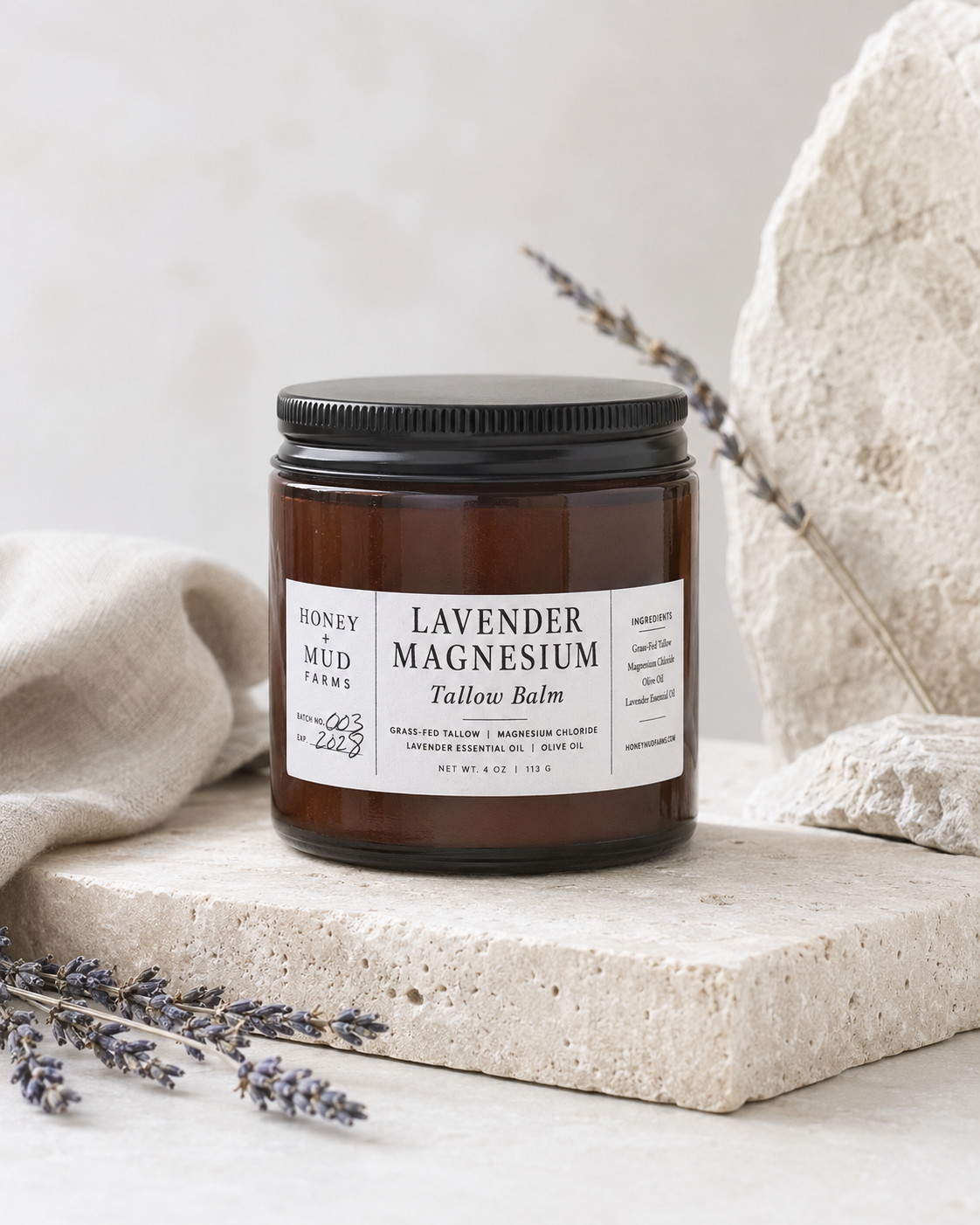A jar of Honey Mud Farms Lavender Magnesium Tallow Balm sits on a light stone slab with lavender sprigs and neutral fabric props against a soft beige background.