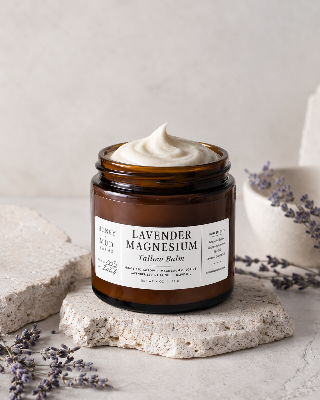 An amber glass jar of Honey Mud Labs Lavender Magnesium Tallow Balm with a white cream product visible at the top, styled on textured stone slabs with lavender sprigs against a neutral light beige background.