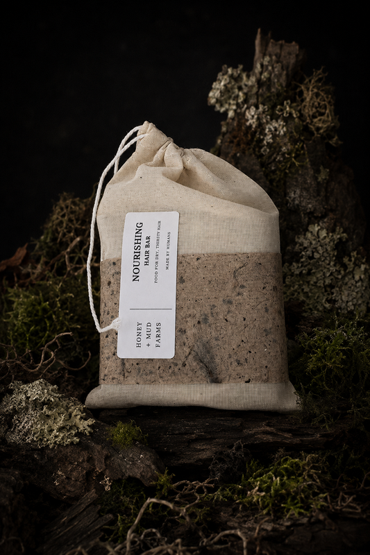 A drawstring fabric pouch containing honeycomb blocks, labeled nourishing hair bar, styled on a mossy log against a dark background.