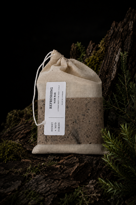 A small natural linen drawstring pouch with a white label reading 'Refreshing Hair Bar' placed on a dark forest floor among moss, bark, and evergreen branches.