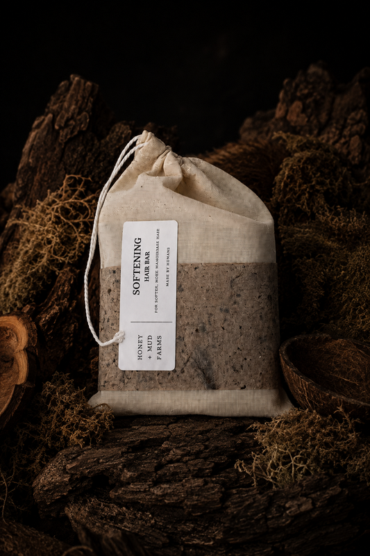 A drawstring fabric pouch containing a dark brown herbal or tea-like blend, with a white label reading 'SOFTENING' and smaller text 'HONEY FARM', styled on dark wood and moss against a black background.