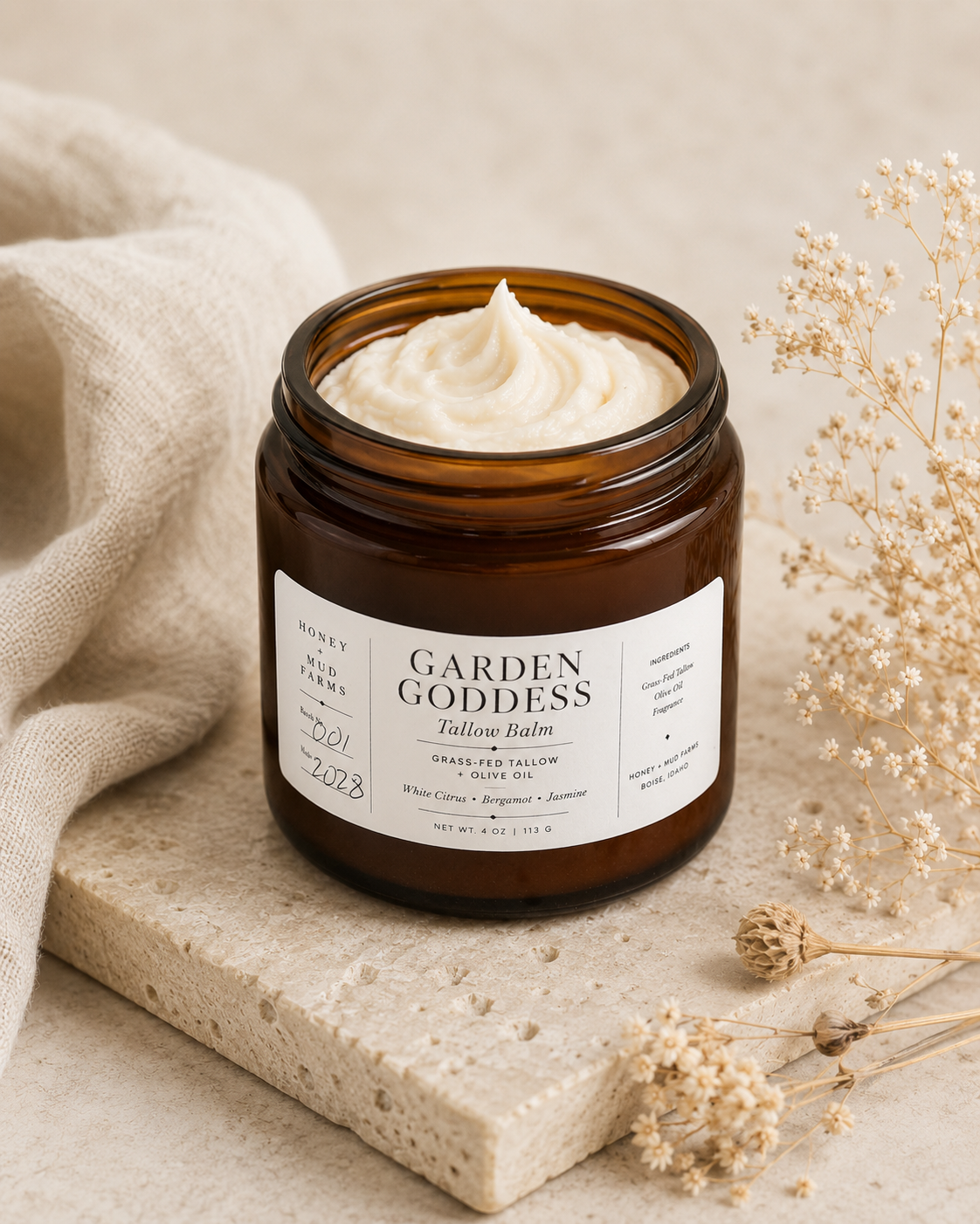 An amber glass jar candle filled with whipped cream-colored balm, labeled 'GARDEN GODDESS Talloo Balm', styled on a stone slab with a linen cloth and dried flowers.